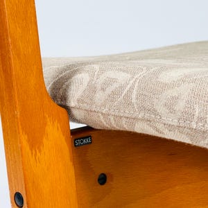 Pode incluir: Close-up de uma cadeira alta de madeira com uma almofada bege estampada. A cadeira tem um acabamento castanho claro e uma pequena etiqueta "STOKKE". A almofada tem um design floral subtil.