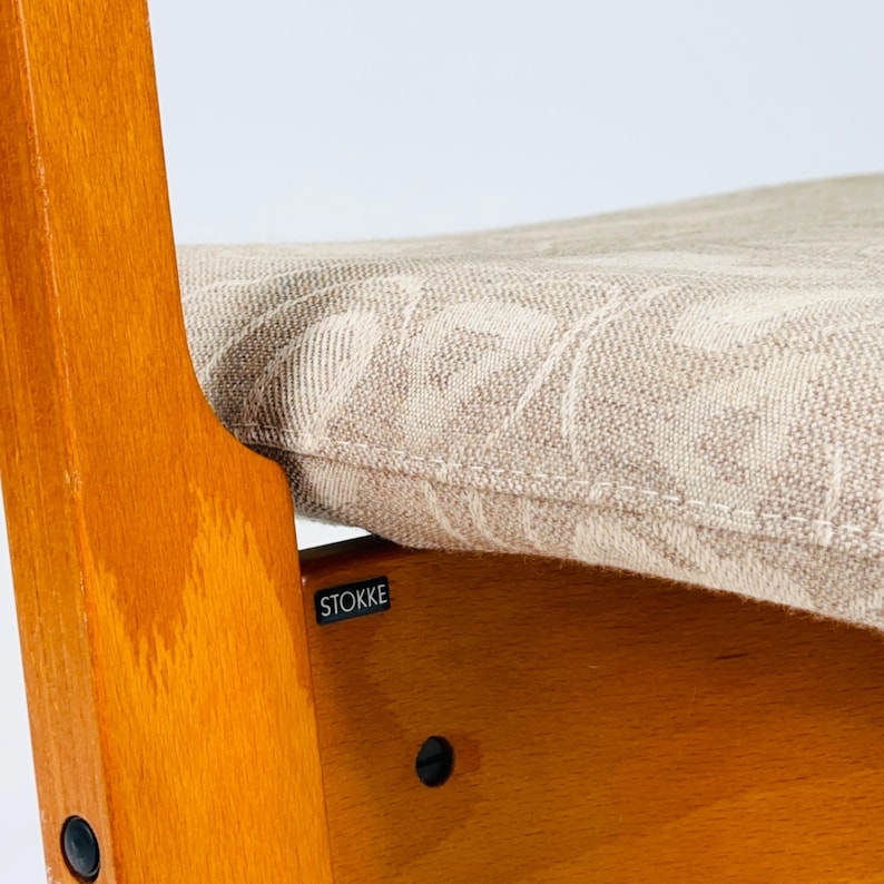 Pode incluir: Close-up de uma cadeira alta de madeira com uma almofada bege estampada. A cadeira tem um acabamento castanho claro e uma pequena etiqueta "STOKKE". A almofada tem um design floral subtil.
