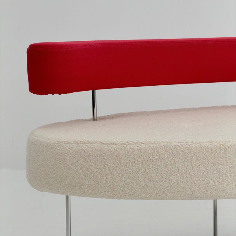 K&ouml;nnte beinhalten: Ein Sofa mit einer cremefarbenen, strukturierten Sitzfl&auml;che in ovaler Form im Boucl&eacute;-Stil. Eine lange, horizontale, rote R&uuml;ckenlehne ist &uuml;ber der Sitzfl&auml;che angebracht und wird von d&uuml;nnen, vertikalen Stangen aus poliertem Chrommetall gest&uuml;tzt.