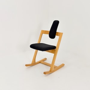 May include: A wooden chair with a black seat and backrest. The chair has a unique design with angled legs and a curved back support. The frame is made of light-colored wood, and the chair is set against a white background.