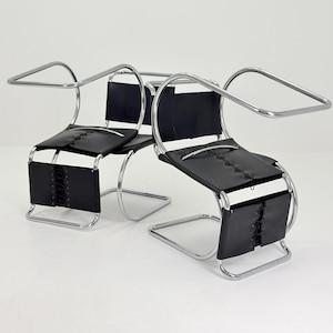 May include: Two chairs featuring a continuous tubular chrome frame that forms the armrests and base. The seat and backrest consist of black leather panels secured with visible lacing details along the center of the seat and the front panel.