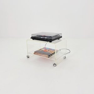 May include: A clear acrylic record player stand with a black record player on top and a stack of vinyl records on the bottom shelf. The stand has small wheels for easy movement.