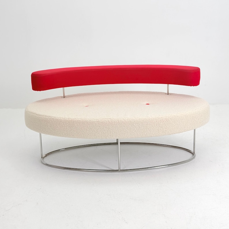 K&ouml;nnte beinhalten: Ein ovales Sofa mit einer cremefarbenen, strukturierten Sitzfl&auml;che mit zwei getufteten Kn&ouml;pfen und einer geschwungenen, leuchtend roten R&uuml;ckenlehne. Das M&ouml;belst&uuml;ck wird von einem d&uuml;nnen, silberfarbenen Metallrahmen getragen, der eine ovale Basis bildet.