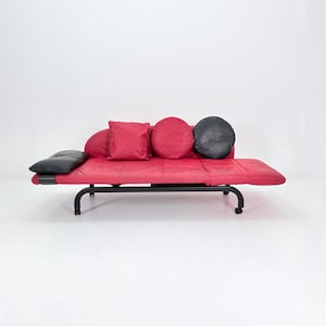 May include: A red leather sofa with a black metal frame and several cushions. The sofa has a rectangular shape with a quilted surface. The cushions are red and black, with square and round shapes. The sofa is on wheels.