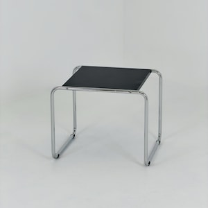 May include: A square side table featuring a smooth black tabletop surface supported by a continuous, tubular chrome-plated metal frame. The frame forms a U-shaped base on each side, providing a stable structure for the flat, dark-colored top.
