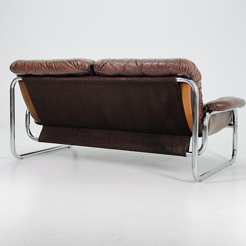 May include: A brown leather two-seater sofa with a chrome frame. The sofa has a tufted back and a loose cushion.