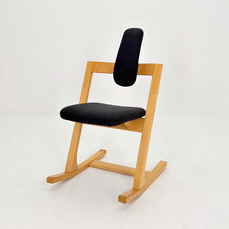 May include: A wooden rocking chair with a black seat and backrest. The chair has a light-colored wooden frame and a curved base. The backrest is a padded, vertical rectangle. The seat is a padded, horizontal rectangle.