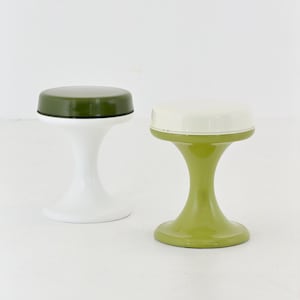 May include: Two hourglass-shaped stools with smooth, glossy finishes. The left stool features a dark olive green circular seat atop a white base. The right stool has a cream-colored circular seat supported by a light lime green hourglass base.