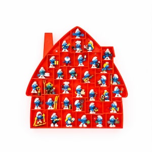 May include: A bright orange house-shaped plastic display shelf with multiple compartments, each containing a small blue Smurf figurine. The collection features various characters in different poses, holding items like musical instruments, tools, and flowers.