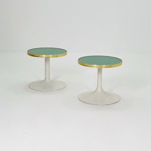 May include: Two small, round side tables. Each table has a light green top with a gold-colored trim. The tables have a white, pedestal-style base.