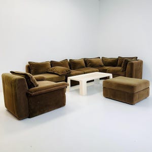 May include: A brown sectional sofa with matching armchair and ottoman. A rectangular, off-white coffee table sits in front of the sofa. The furniture has a plush, velvet-like texture. The room has white walls and a white floor.