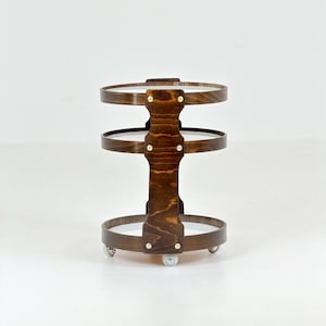 May include: A three-tiered circular rolling cart constructed from dark stained wood with visible grain. The structure features a central vertical support column connected to three round shelves, accented with small brass-colored metal screws.