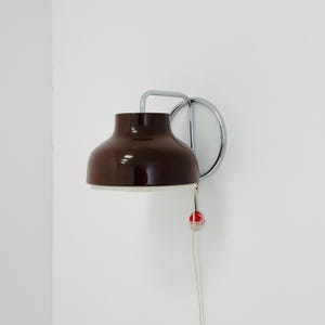 Puede incluir: Una lámpara de pared marrón y blanca con un marco de metal cromado. La lámpara tiene una pantalla redondeada y un cordón con un acento rojo. La lámpara está montada en una pared blanca.