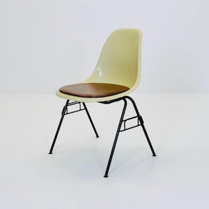 May include: A retro-style chair featuring a pale yellow plastic seat and a brown leather cushion. It has a black metal frame with four legs. The chair's design is minimalist and would suit a kitchen or study setting.
