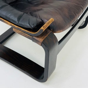 May include: Close-up of a dark brown leather chair with a wooden frame. The chair has a curved, rectangular base and a padded seat. The leather is worn and has a distressed look.