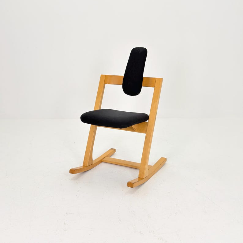 May include: A wooden kneeling chair with a black seat and backrest. The chair has a light wood frame and a curved base. The backrest is a padded, vertical rectangle. The seat is a padded, horizontal rectangle.