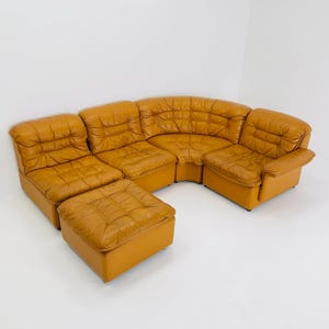 Op de afbeelding: Een modulaire hoekbank en poef set in mid-century modern stijl. De set heeft een warme, goudbruine lederen bekleding met een gecapitonneerd ontwerp. De hoekbank is gebogen om een afgeronde vorm te creëren en de poef is vierkant.
