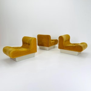 Op de afbeelding: Drie mosterdgele fluwelen fauteuils met een afgerond ontwerp en knoopversteviging. De stoelen staan op een witte basis. De stoelen zijn in een rij op een witte achtergrond geplaatst.