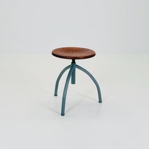 Pode incluir: Um pequeno banco redondo de madeira com assento castanho e três pernas azuis curvas. O banco tem um design minimalista. O assento tem cerca de 30 cm de diâmetro. Adequado para várias configurações.