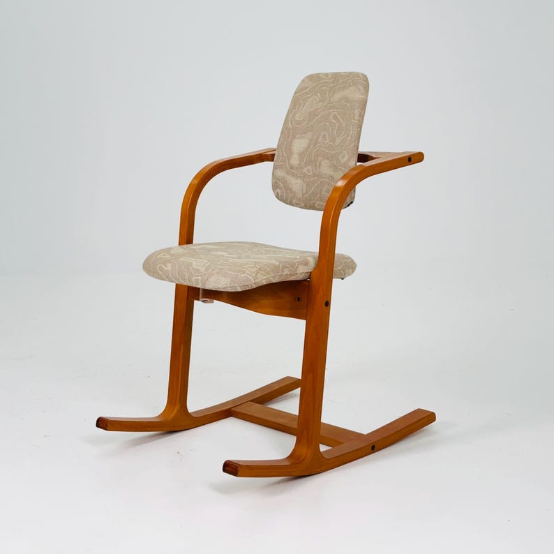 Pode incluir: Cadeira de baloi&ccedil;o de madeira com acabamento castanho claro. A cadeira apresenta encosto e apoios de bra&ccedil;os curvos, e o assento e o encosto s&atilde;o estofados em tecido bege estampado. A cadeira tem um design cl&aacute;ssico e moderno de meados do s&eacute;culo.