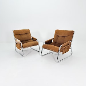 May include: A pair of mid-century armchairs with chrome frames and brown suede cushions. Each chair has a side pocket. One pocket contains a magazine. The chairs are a stylish design.