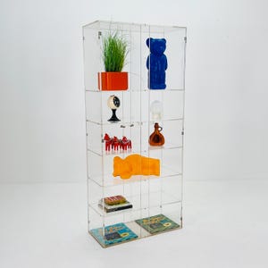 May include: A clear acrylic display cabinet with multiple shelves, showcasing various decorative items. The items include a blue bear figurine, a potted plant, a small clock, and other colorful ornaments. The cabinet is against a white background.