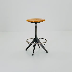 Pode incluir: Banqueta ajustável de estilo industrial vintage com assento redondo de madeira castanha clara. A base de metal é cinza escuro com apoio para os pés e design de três pernas. A altura da banqueta é ajustável.