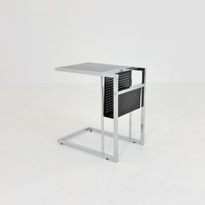 以下が含まれることがあります： 光沢のあるクロームメッキの金属フレームとダークグレーの正方形の天板を備えたC字型のサイドテーブル。テーブルの側面には、本や雑誌を収納するための穴あきグリッドパターンの黒い金属製マガジンラックが付いています。