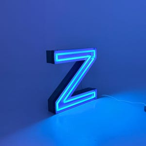 May include: A neon sign in the shape of the letter "Z". The sign is outlined with bright blue neon lights, encased in a black frame. The background is a solid blue color, creating a striking contrast.