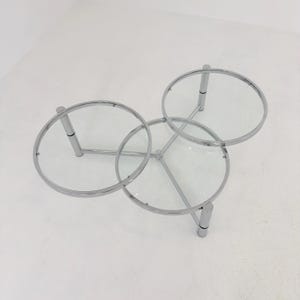 May include: A coffee table featuring three circular clear glass tops supported by a polished chrome metal frame. The structure consists of interconnected tubular legs and rings that hold each glass surface at a consistent height.