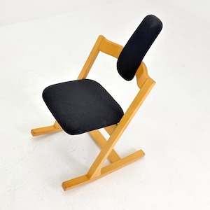 May include: A wooden kneeling chair with a black seat and backrest. The chair has a light wood frame and a minimalist design. The seat and backrest are made of a dark fabric. The chair is designed to promote good posture.
