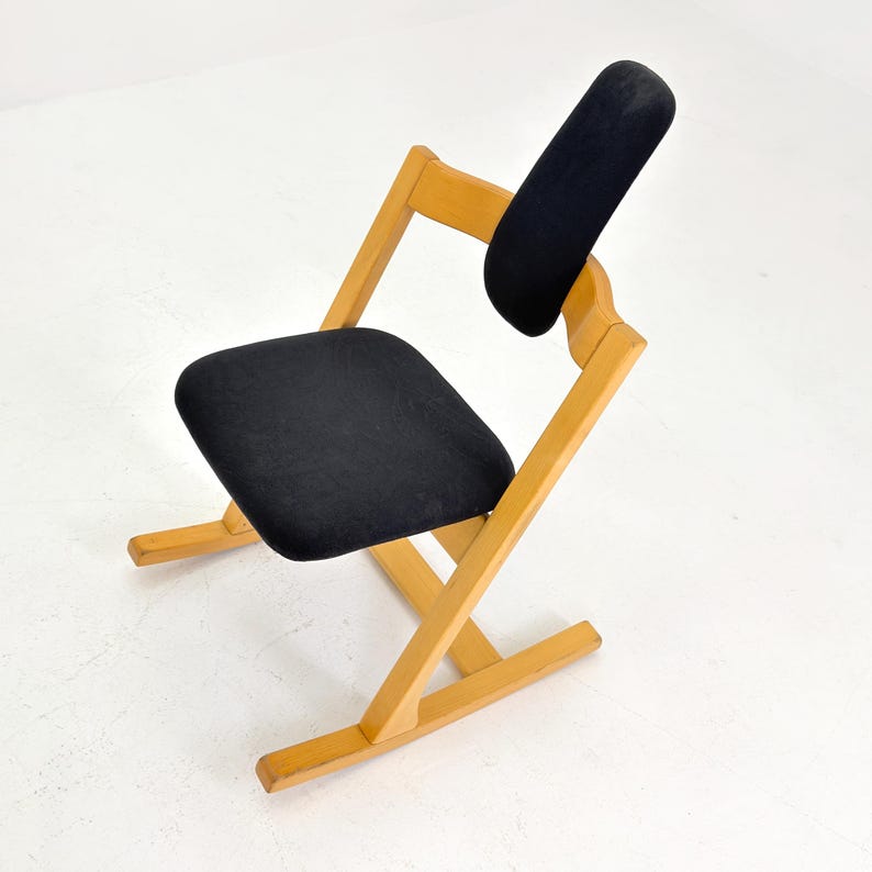 May include: A wooden kneeling chair with a black seat and backrest. The chair has a light wood frame and a minimalist design. The seat and backrest are made of a dark fabric. The chair is designed to promote good posture.