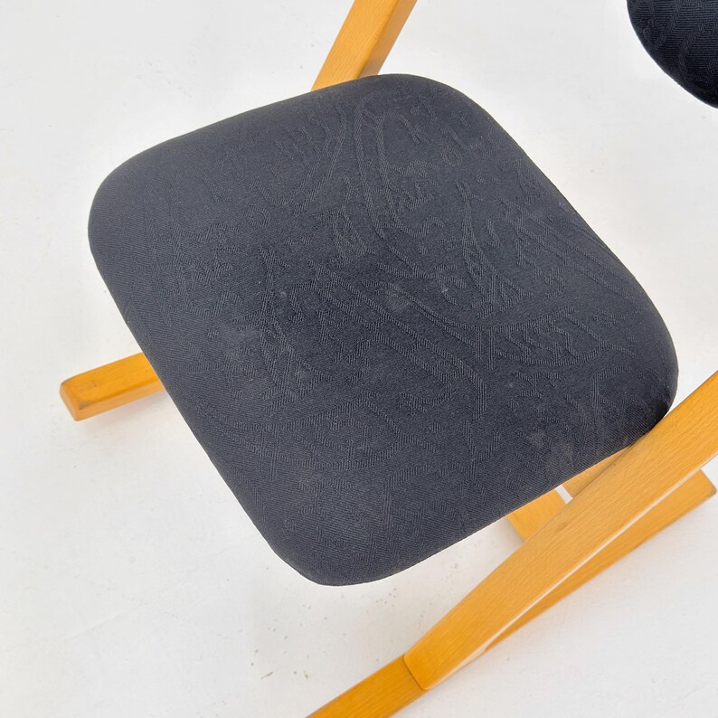 May include: Close-up of a black upholstered chair seat with a subtle pattern. The seat is supported by a light-colored wooden frame. The chair is likely part of a set, with a simple, functional design.