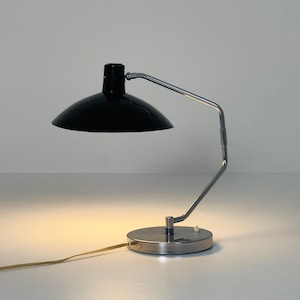 May include: A chrome and black desk lamp with a round base and a flexible arm. The lamp has a black shade and a white switch.