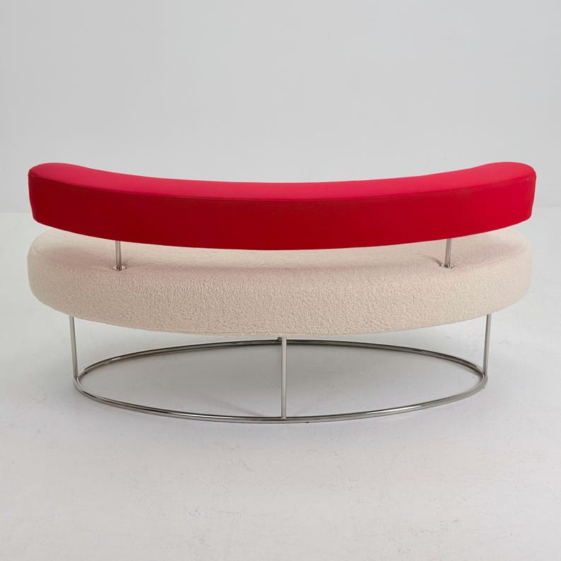 K&ouml;nnte beinhalten: Ein geschwungenes Sofa mit einer cremefarbenen, strukturierten Sitzfl&auml;che und einer leuchtend roten R&uuml;ckenlehne. Die R&uuml;ckenlehne wird von zwei d&uuml;nnen Metallstangen gest&uuml;tzt und das gesamte M&ouml;belst&uuml;ck ruht auf einem ovalen Metallgestell.