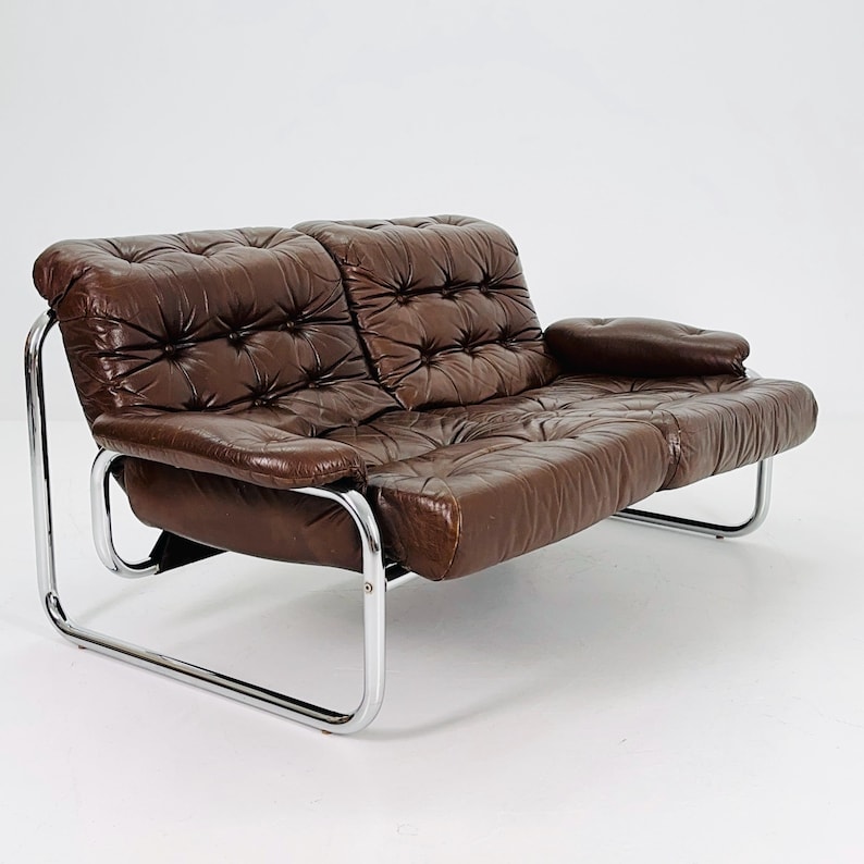 May include: A brown leather two-seater sofa with a chrome frame. The sofa has a tufted design and a relaxed, comfortable look.
