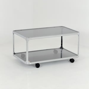 Peut inclure: Une desserte rectangulaire à deux niveaux dotée d'une structure en métal chromé poli aux coins arrondis. La desserte comprend deux étagères en verre fumé foncé et repose sur quatre roulettes pivotantes noires fixées à la base.