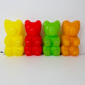 Könnte beinhalten: Vier große, farbenfrohe Gummibär-förmige Dekorationen in Gelb, Rot, Grün und Orange. Jeder Bär hat eine glatte, glänzende Oberfläche und ein klassisches Gummibär-Design. Sie stehen aufrecht vor einem weißen Hintergrund.