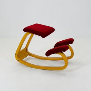 Puede incluir: Una silla de rodillas con una estructura de madera clara y curvada. Cuenta con un asiento rectangular rojo y dos rodilleras rojas. La silla está diseñada para promover una buena postura y soporte ergonómico.