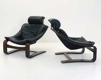 May include: Two black leather lounge chairs with dark wooden frames. The chairs have a curved design with headrests and armrests. The leather appears worn, and the wood has a dark finish. The chairs are set against a white background.
