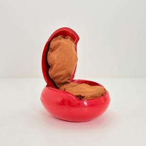 Op de afbeelding: Een felrode, retro stoel met een afgeronde basis en een open, eivormige bovenkant. De binnenkant heeft een zacht, bruin kussen. De stoel is gemaakt van glanzend plastic en staat tegen een effen witte achtergrond.