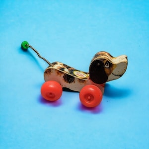 Op de afbeelding: Een vintage houten speelgoedhond met bruine en zwarte vlekken. De hond heeft rode wielen en een trekkoord. Het gezicht van de hond is geschilderd met bruine ogen en een zwarte neus. De hond heeft een label met de tekst "Tiny Tot" erop.