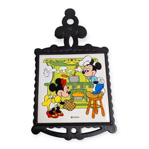 May include: Black metal trivet featuring a colorful image of Mickey and Minnie Mouse cooking in a kitchen. The image is a ceramic tile with a scalloped black metal frame and a decorative top. The Disney logo is visible.