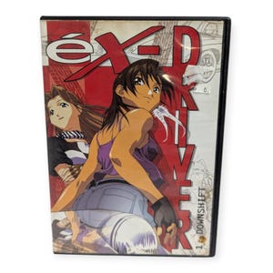 May include: DVD case with anime-style artwork. The cover displays the title "éX-Driver" in white and black against a red backdrop. Two female characters are shown, one in a purple top and the other in a black top. The text "1 DOWNSHIFT" is also visible.
