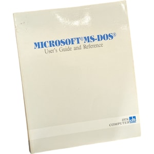 Op de afbeelding: Een wit boek met de titel "MICROSOFT® MS-DOS® Gebruikershandleiding en Referentie" in blauw gedrukt. Het boek heeft een blauw en wit DTK COMPUTER logo in de rechterbenedenhoek.