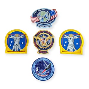 Könnte beinhalten: Fünf gestickte Patches mit Motiven von Space-Shuttle-Missionen. Die Patches sind rund und rechteckig und in den Farben Blau, Rot, Weiß und Gelb gehalten. Die Patches zeigen Astronauten, die Erde und die amerikanische Flagge.