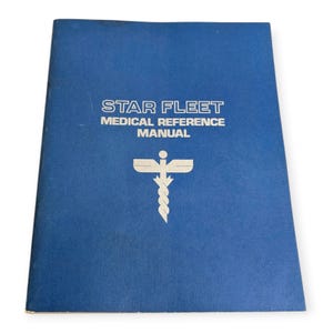 May include: A blue book titled "STAR FLEET MEDICAL REFERENCE MANUAL" with white text and a medical symbol. The cover is a solid blue color with some wear and tear. The medical symbol is a caduceus with a cross.