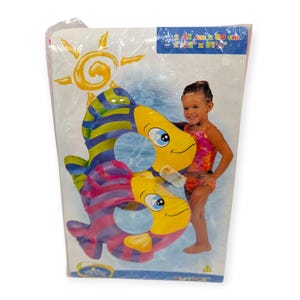 Puede incluir: Un flotador inflable con forma de pez en un paquete de plástico transparente. El flotador tiene rayas amarillas, azules, verdes y rosas. El paquete tiene las dimensiones de 80 cm x 80 cm y muestra a una niña en traje de baño.