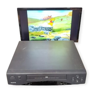 Peut inclure: Un lecteur VHS Sanyo noir avec une fente de chargement frontale, un bouton d'alimentation, un bouton d'éjection et des commandes de lecture. Derrière le lecteur, un écran plat affiche une scène animée colorée avec un ours jaune dans un paysage.