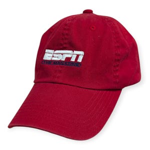 May include: A red baseball cap with the ESPN logo embroidered on the front. The logo is white with a blue outline and the text "THE MAGAZINE" underneath. The cap has a curved brim and a button on top.